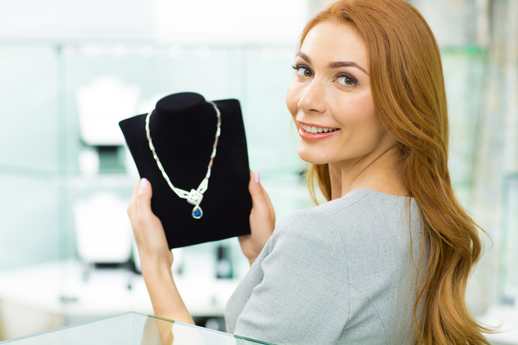 Gorgeous happy woman smiling to the camera over her shoulder holding beautiful diamond necklace choosing jewelry at the store consumptions retail money consumerism customer client elegance.